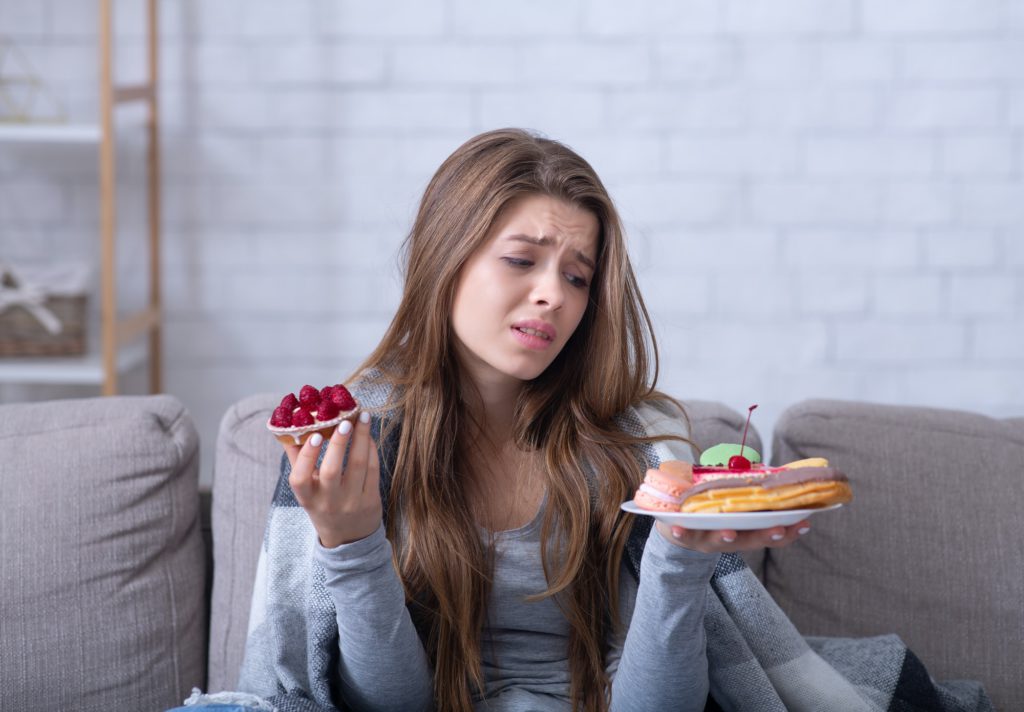 Depressed young lady having eating disorder, comforting herself with sweets on sofa at home