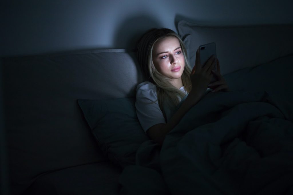 Social networks addiction, insomnia. Young woman using smartphone, lying on bed at late night.