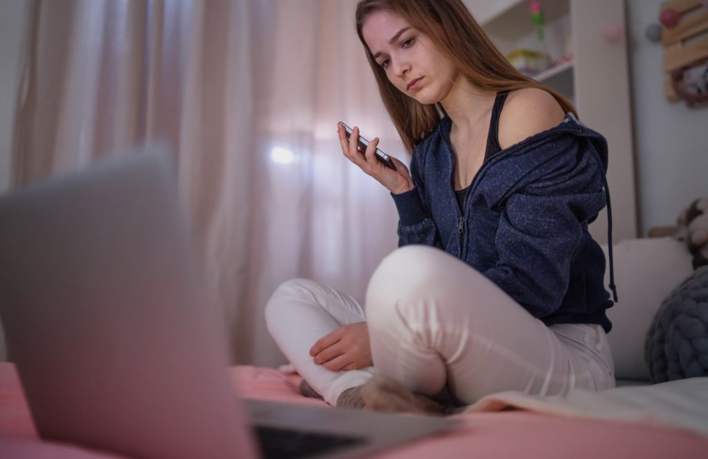 Worried young girl with smartphone sitting indoors on bed, online dating concept